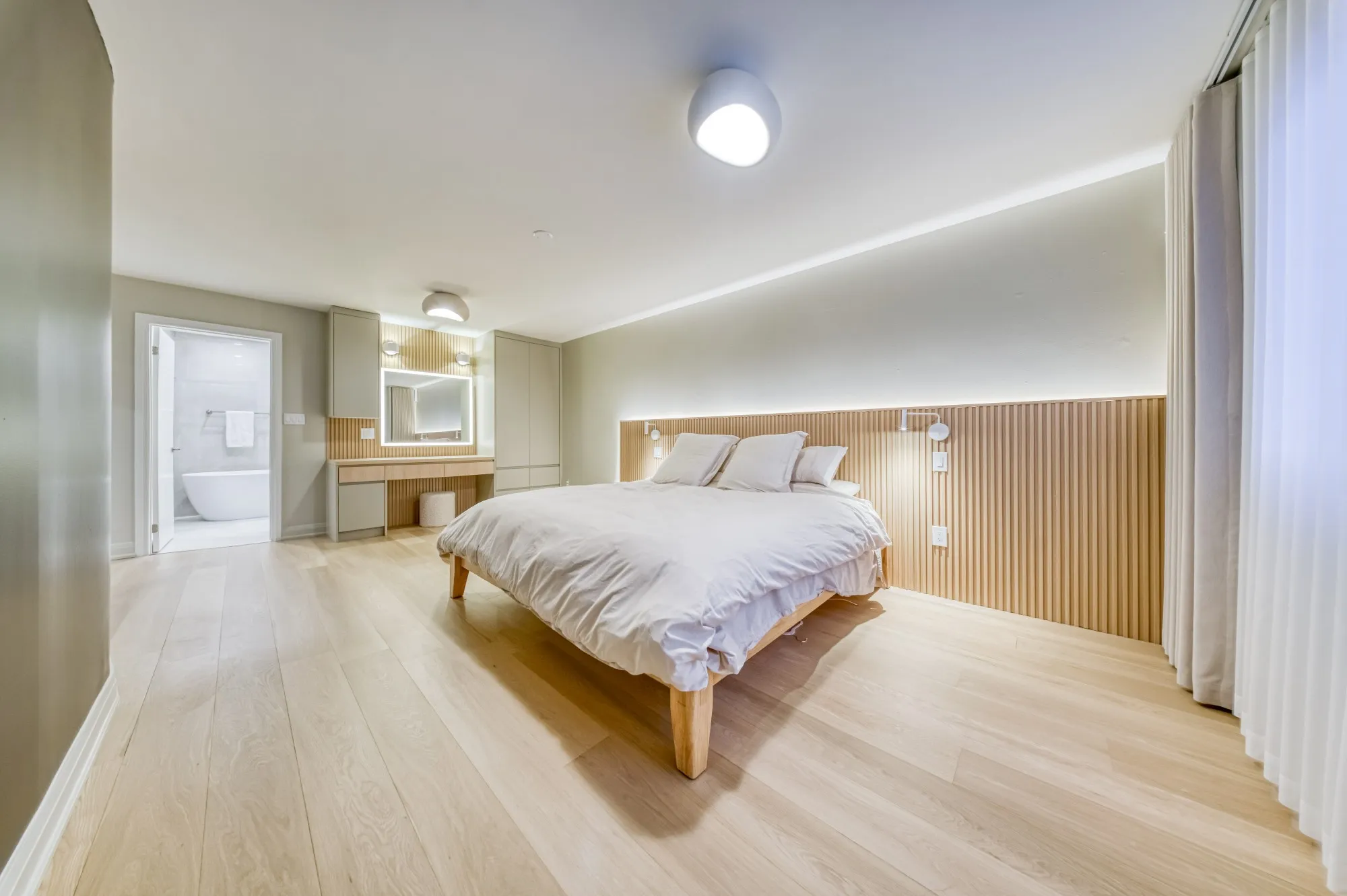 A primary bedroom with a slatted oak wall behind the bed, wide plank floors and a lit vanity, with the ensuite beyond.