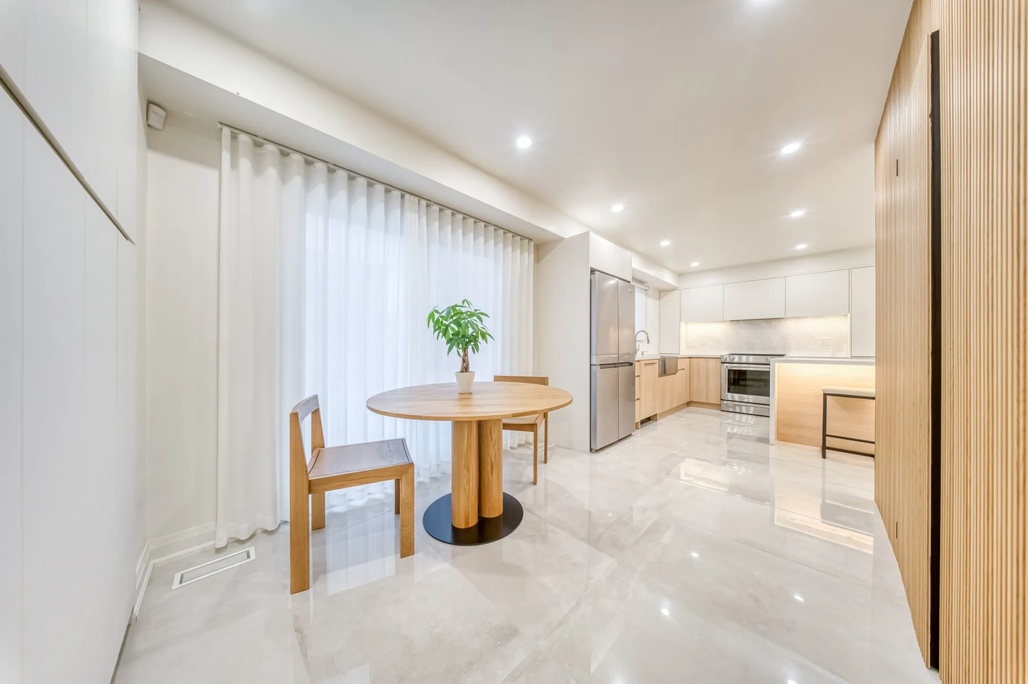 A round oak table by sheer curtains, a slatted oak wall and the kitchen beyond.