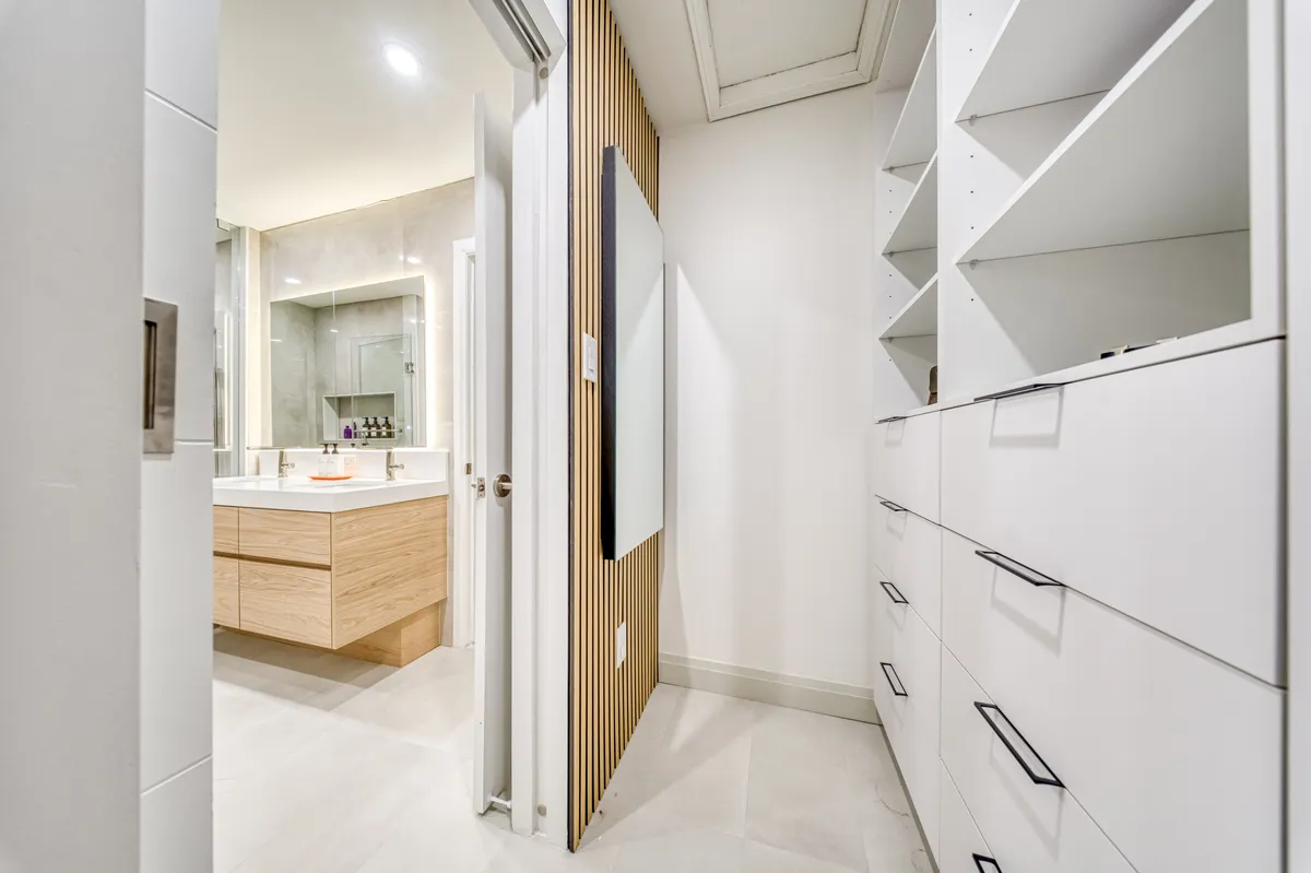 A walk-in closet in white with open shelving, drawers and a slatted oak panel, with the ensuite beyond.